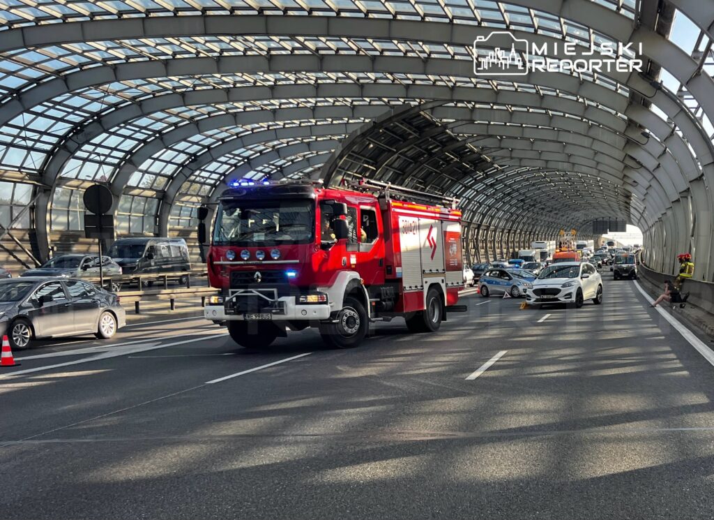 Czerwony wóz strażacki przejeżdża przez tunel, w tle zablokowany ruch samochodowy i osoby w mundurach.