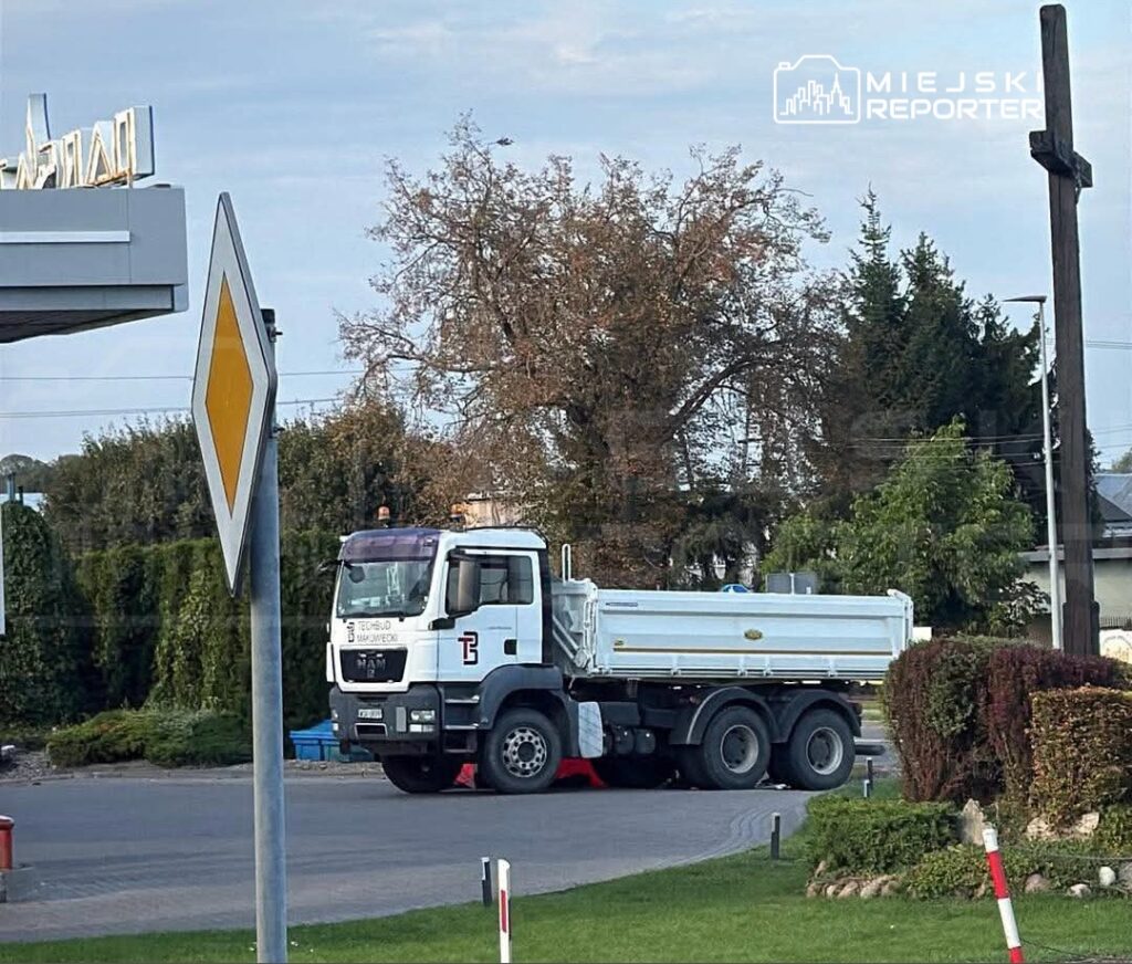 Ciężarówka z białą skrzynią ładunkową stoi na skrzyżowaniu, otoczona drzewami i ogrodzeniem.