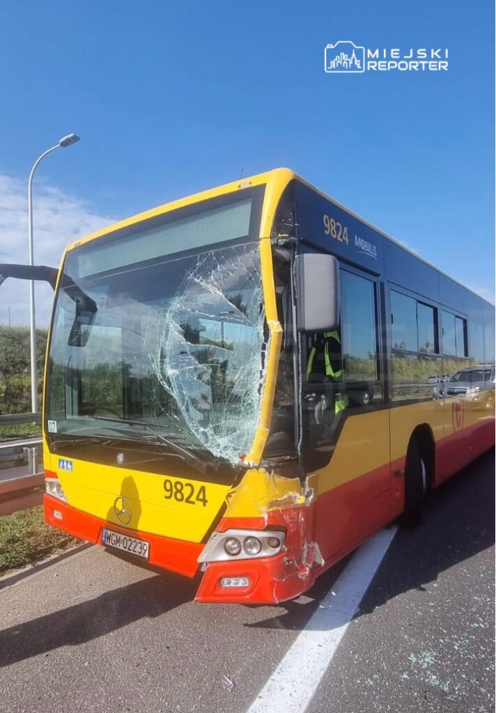Uszkodzony autobus z pękniętą szybą czołową stoi na pasie drogowym, obok widać barierki ochronne i zieleń.