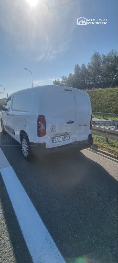 Biały samochód dostawczy zaparkowany na poboczu drogi, obok barierki ochronnej, w słoneczny dzień.
