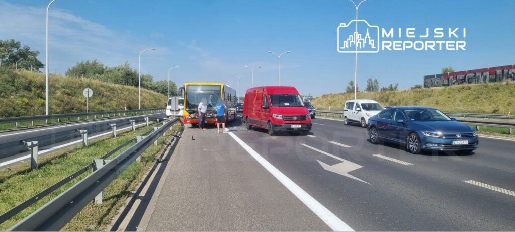 Na drodze znajduje się uszkodzony autobus oraz kilka innych pojazdów, a obok mężczyźni sprawdzają stan pojazdu.