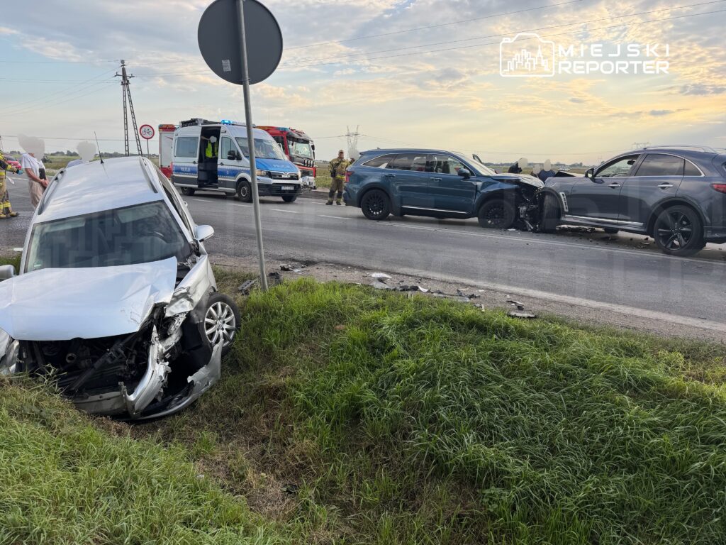 W wyniku wypadku drogowego dwa uszkodzone samochody stoją na poboczu, a obok nich pracują służby ratunkowe.