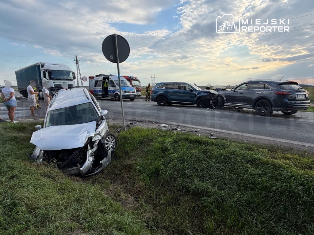 Wielka kolizja drogowa z udziałem trzech pojazdów na drodze, samochód osobowy w rowie, w tle karetka i ciężarówka.