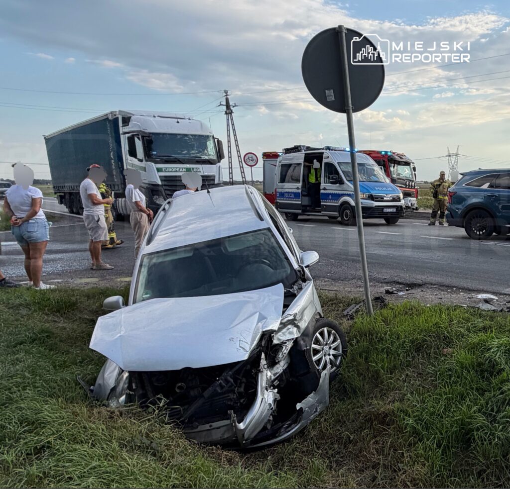 Zniszczony samochód osobowy w rowie przy drodze, obok stoją pojazdy służb ratunkowych i grupka ludzi obserwujących zdarzenie.