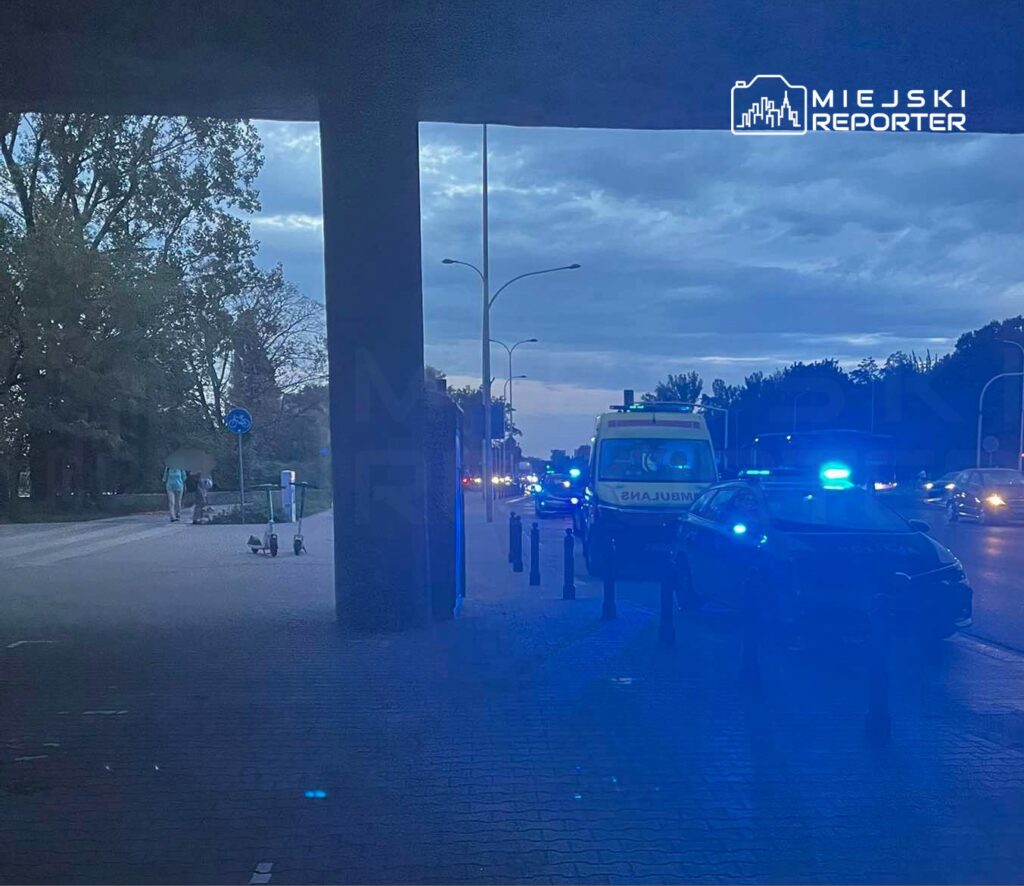 Na chodniku obok ulicy stoją dwa skutery, a w tle widoczne są pojazdy policyjne i karetka z włączonymi sygnałami świetlnymi.