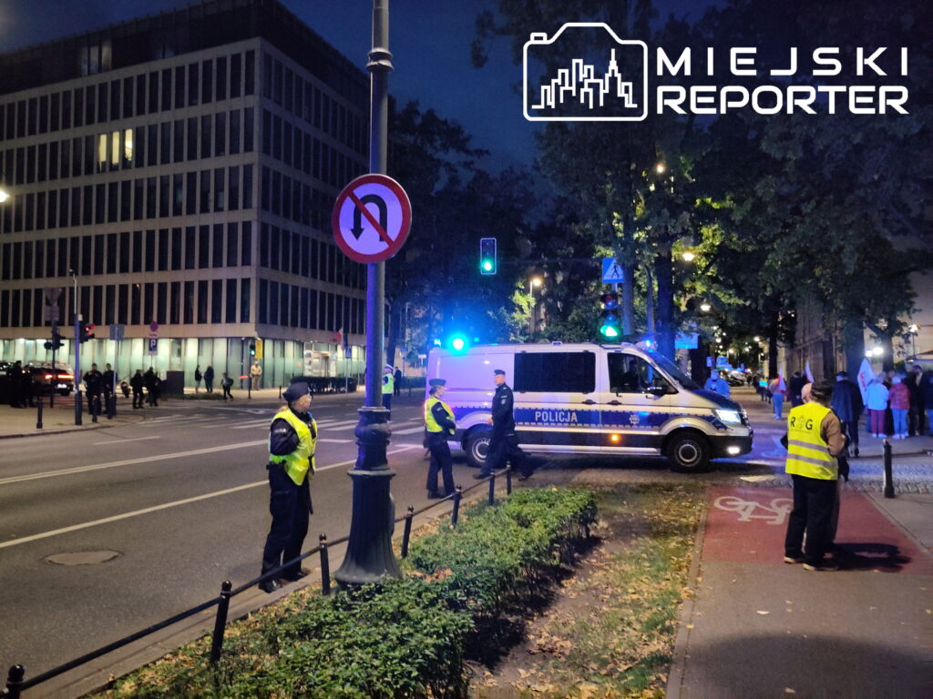 Grupa policjantów w kamizelkach odblaskowych stoi obok policyjnego furgonu na ulicy w mieście, w tle widać ludzi z transparentami.