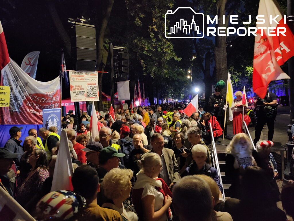 Tłum ludzi z flagami, transparentami i plakatami zgromadzony na ulicy, w tle oświetlone drzewka.