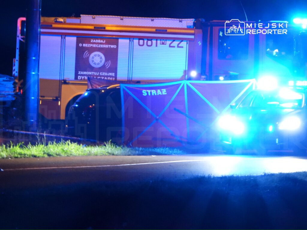 Na drodze stoi samochód policyjny z włączonymi syrenami oraz wóz strażacki z widocznym napisem "Zadbaj o bezpieczeństwo".