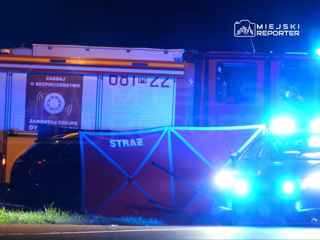 Strażacy przy wozie strażackim z niebieskimi światłami, zabezpieczający teren z czerwonym parawanem z napisem "STRAŻ".
