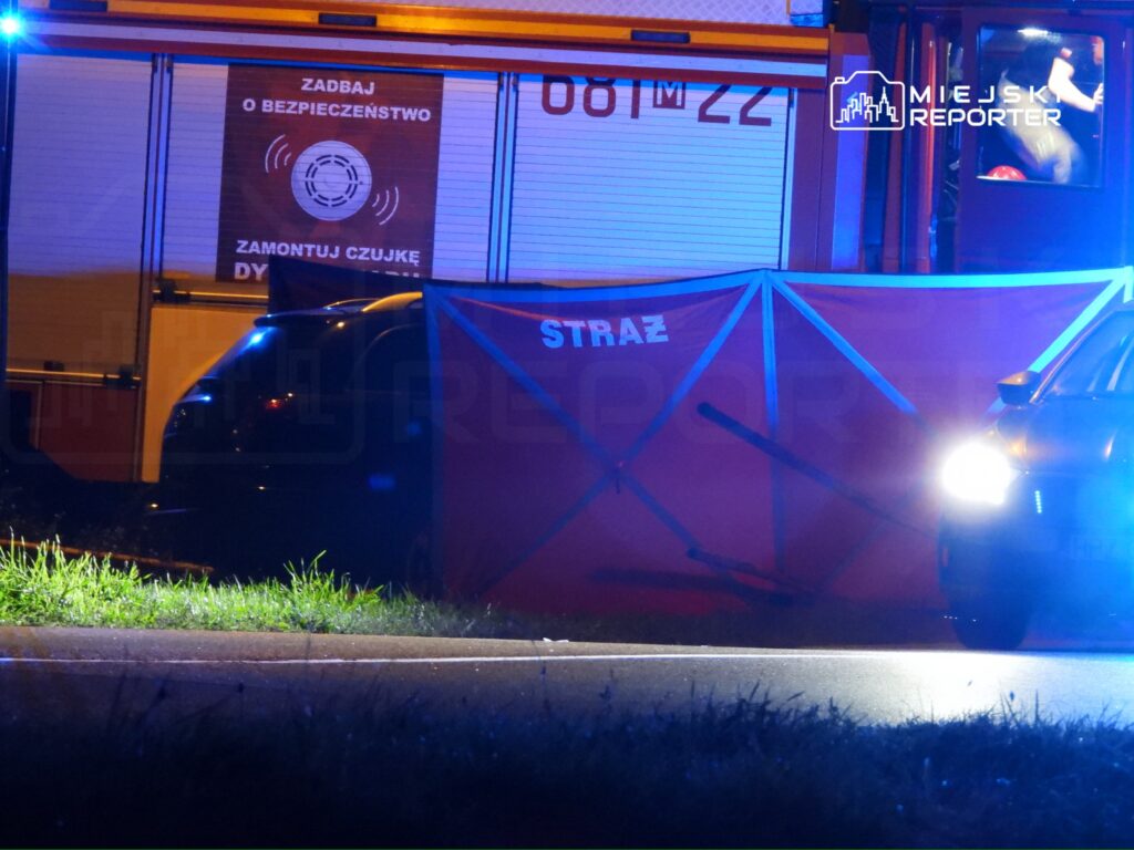Straż pożarna z włączonymi sygnałami świetlnymi zabezpiecza miejsce zdarzenia, a w tle widać czerwony namiot z napisem "STRAŻ".