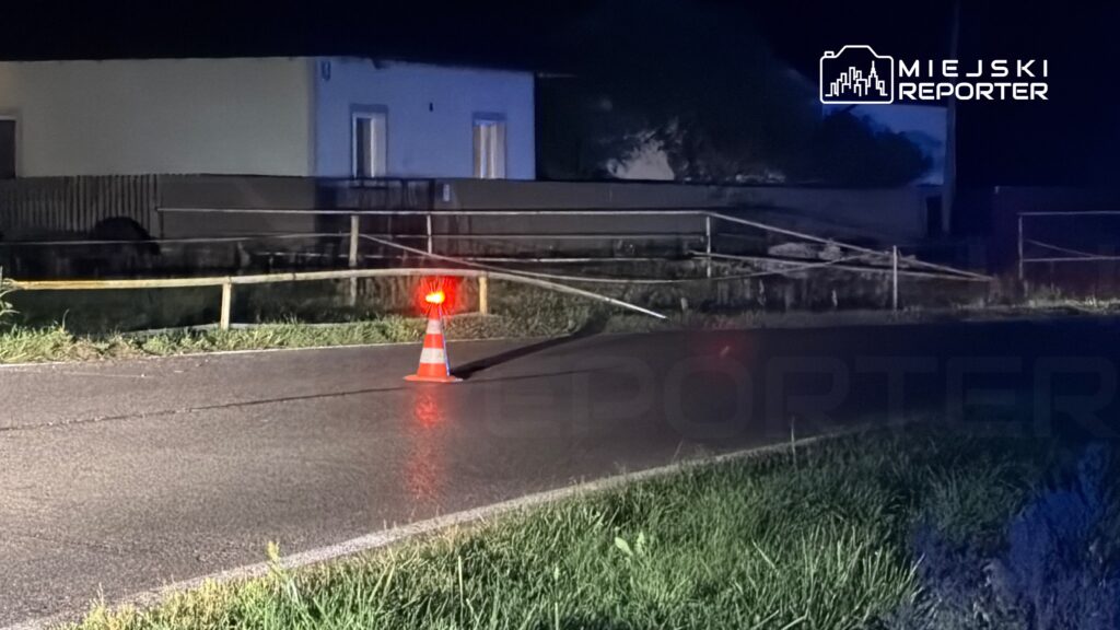 Na drodze znajduje się pachołek drogowy z migającym światłem, a w tle widoczny jest ogrodzony teren z budynkiem.
