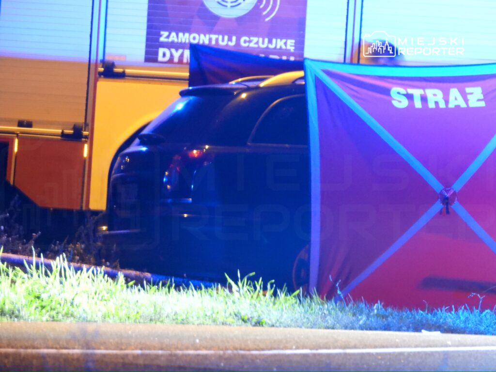 Czarny samochód zaparkowany w pobliżu wozu strażackiego, osłonięty czerwoną płachtą z napisem "STRAŻ".