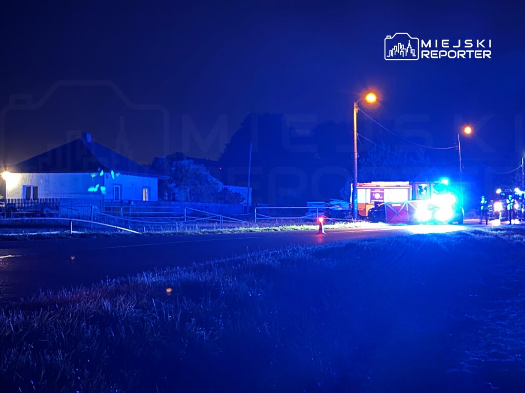 Nocna scena z niebieskimi światłami radiowozu i wozu strażackiego na drodze, obok budynku mieszkalnego.