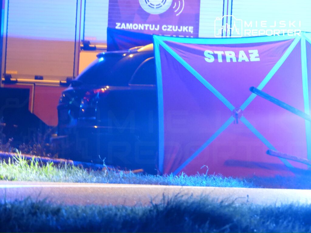 Czarny samochód zaparkowany w pobliżu niebieskiego światła, z namiotem oznaczonym napisem "STRAŻ" w tle.