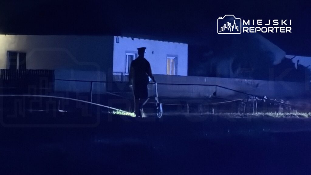 Policjant z latarką przemieszcza się w ciemności obok oświetlonego budynku mieszkalnego.