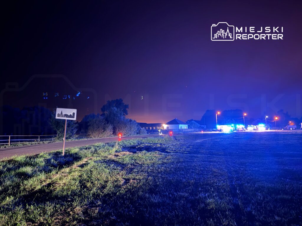 Na drodze wiejskiej widoczny znak drogowy, w tle niebieskie światła pojazdów służb ratunkowych oraz zabudowania.