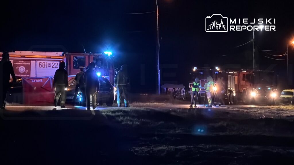 Grupa strażaków i policjantów prowadzi działania ratunkowe w nocy na drodze, otoczeni przez pojazdy służbowe.
