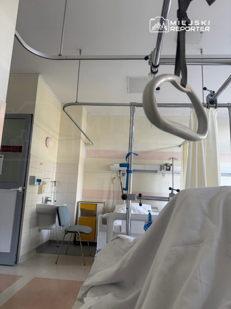 Szpitalna sala z łóżkiem pacjenta, metalowym wózkiem i umywalką, na ścianach jasne kolory.