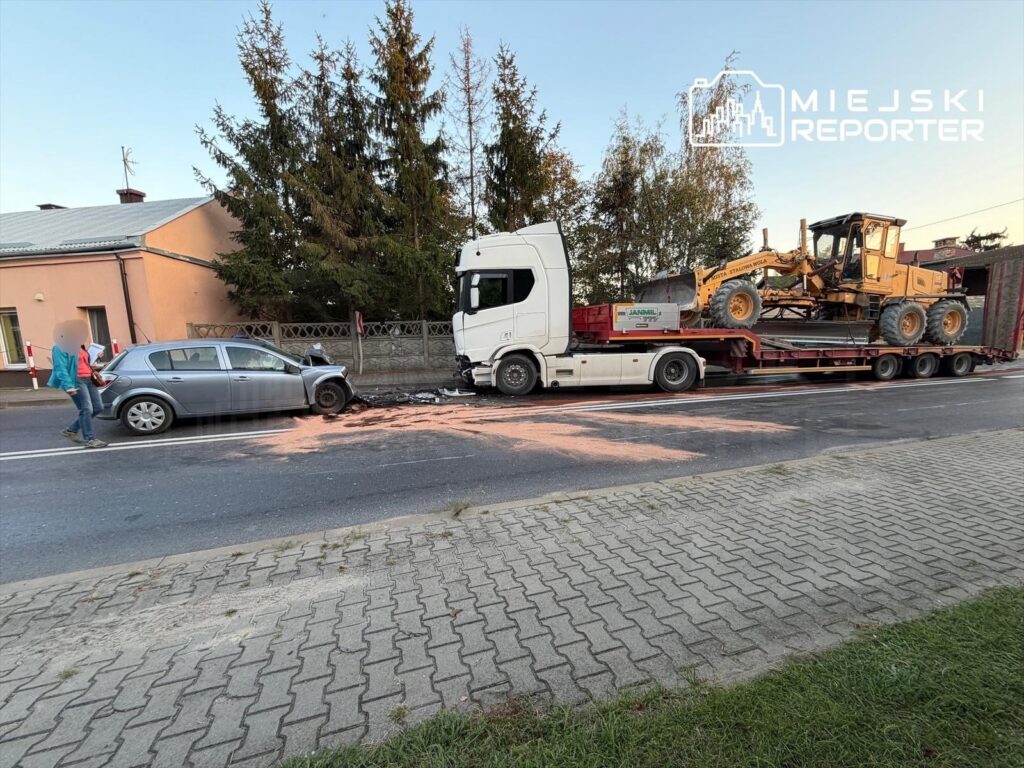 Kolizja drogowa z udziałem szarego samochodu osobowego i ciężarówki transportującej maszynę budowlaną; widoczne uszkodzenia.