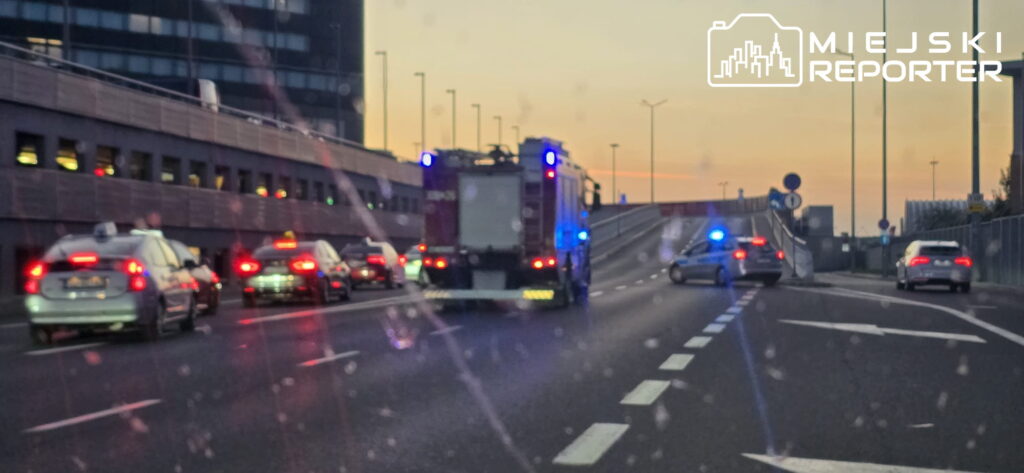 Na drodze przed lotniskiem stoją samochody, w tym radiowóz i wóz strażacki z włączonymi sygnałami świetlnymi.