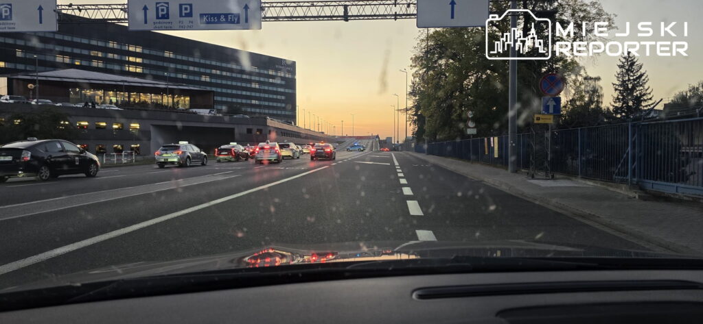 Ruch samochodowy na drodze prowadzącej do lotniska, z widokiem na nowoczesny budynek terminalu w tle.