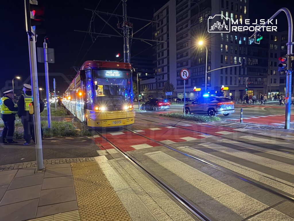 Tramwaj zatrzymany na skrzyżowaniu, obok grupka policjantów rozmawiających z kierowcą, w tle oświetlone budynki.