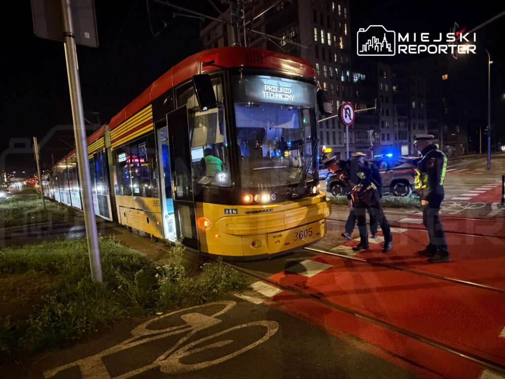 Tramwaj zatrzymany na torach, obok grupy policjantów przeprowadzających kontrolę w nocy.