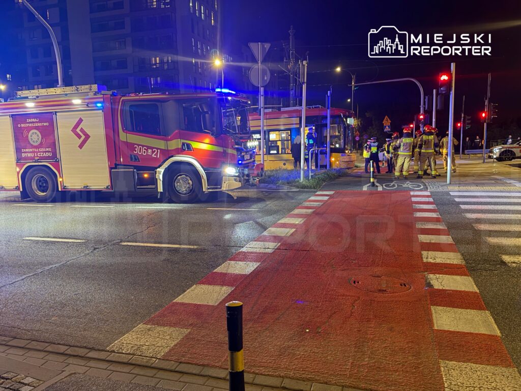 W nocy strażacy w mundurach omawiają sytuację obok wozu strażackiego na skrzyżowaniu z tramwajem.