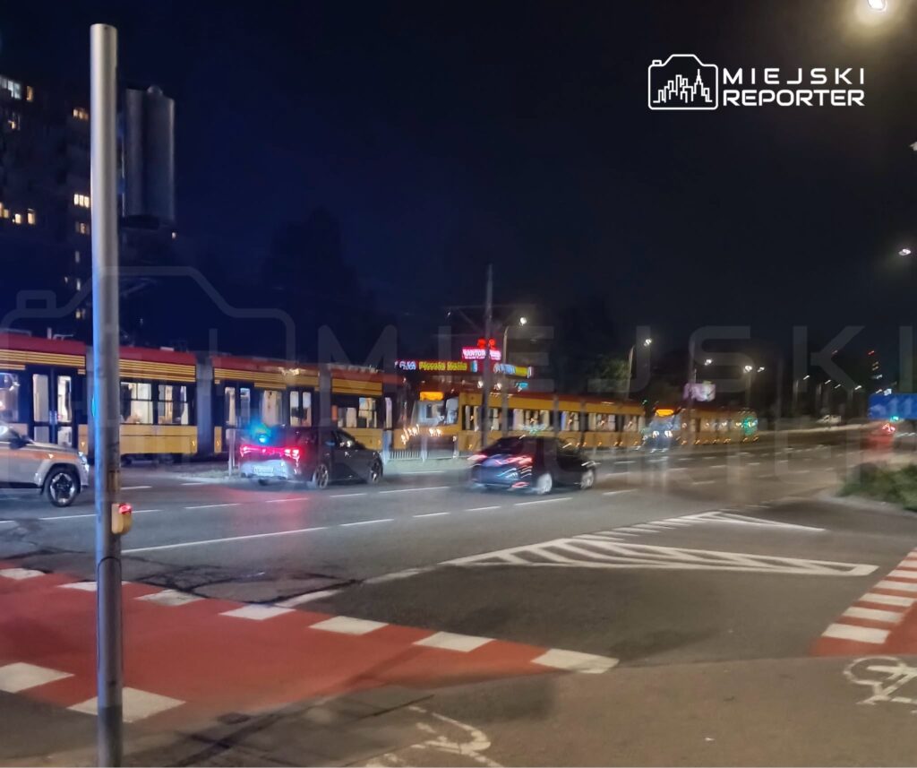 Tramwaj w kolorze żółto-czerwonym zatrzymany na przystanku, obok przejeżdżające samochody w nocy.