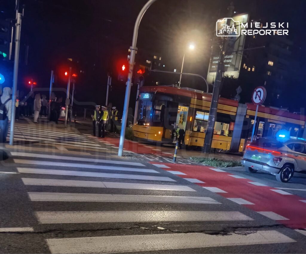 Grupa osób w odblaskowych kamizelkach rozmawia obok tramwaju na czerwonym świetle, w tle widać radiowóz.