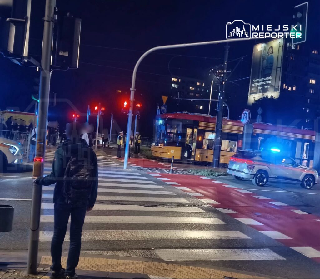 Mężczyzna w plecaku czeka na zielone światło przy przejściu dla pieszych, w tle widać radiowóz i tramwaj.