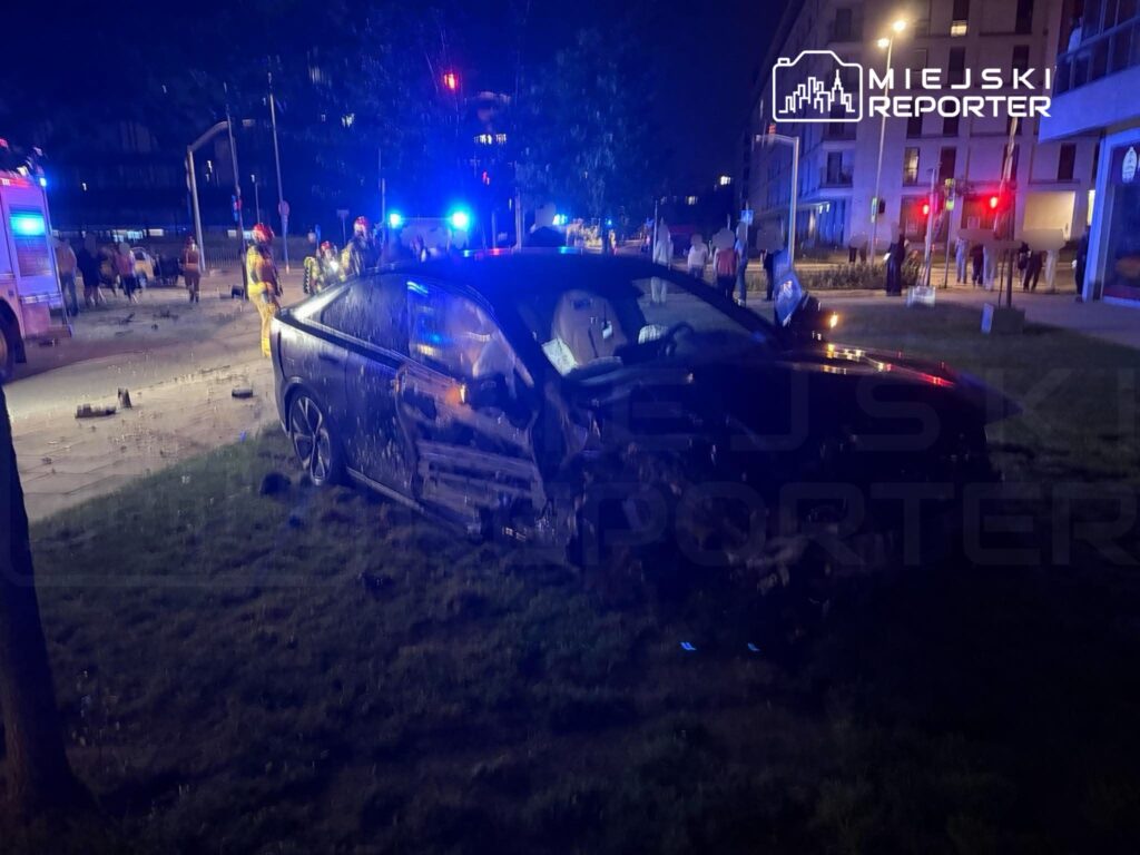 Czarny samochód osobowy z uszkodzoną karoserią stoi na trawie, otoczony przez służby ratunkowe i gapiów w nocy.