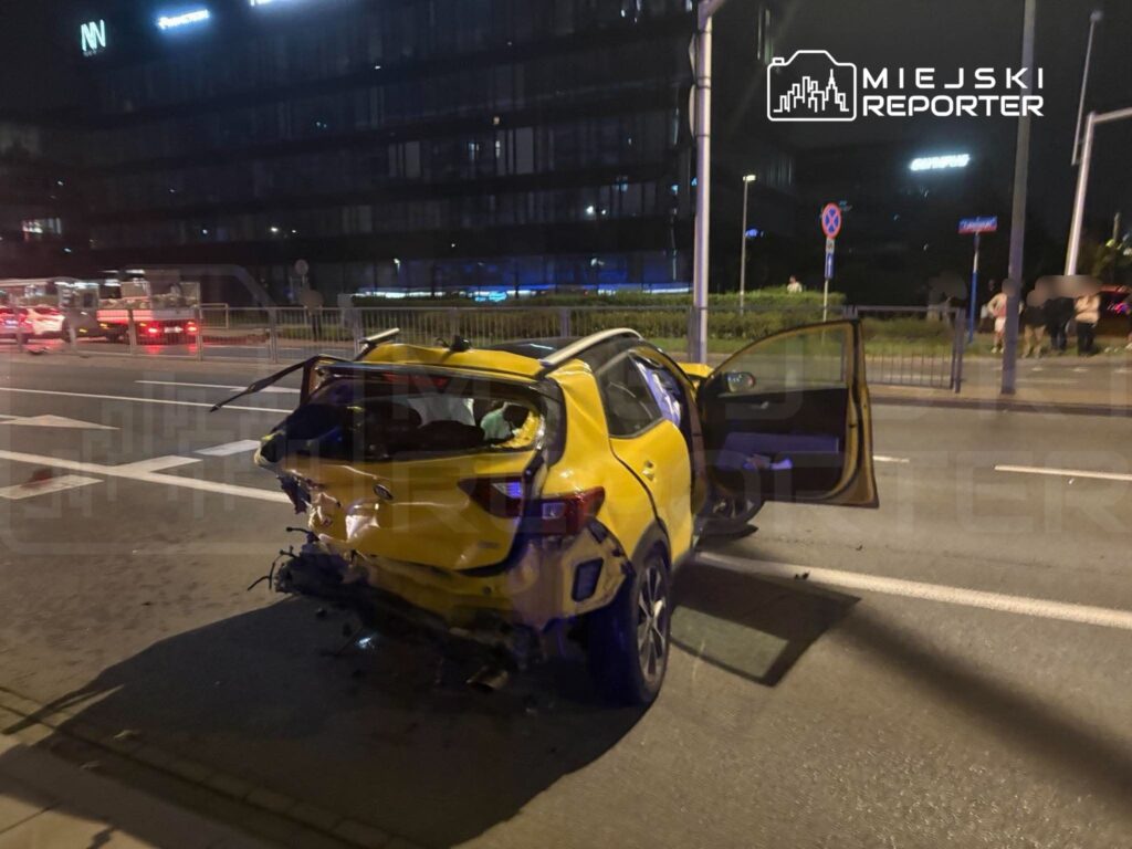 Uszkodzony żółty samochód osobowy stoi na ulicy nocą, z otwartymi drzwiami i widocznymi śladami kolizji.