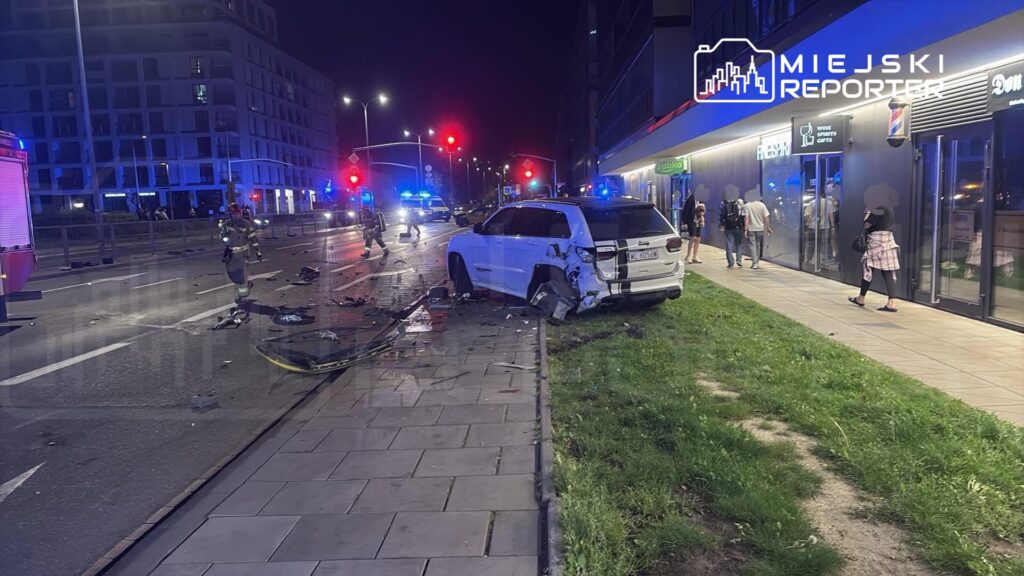 W nocy, na miejskiej ulicy, strażacy pracują przy rozbitym samochodzie osobowym, obok leżą fragmenty wraku.