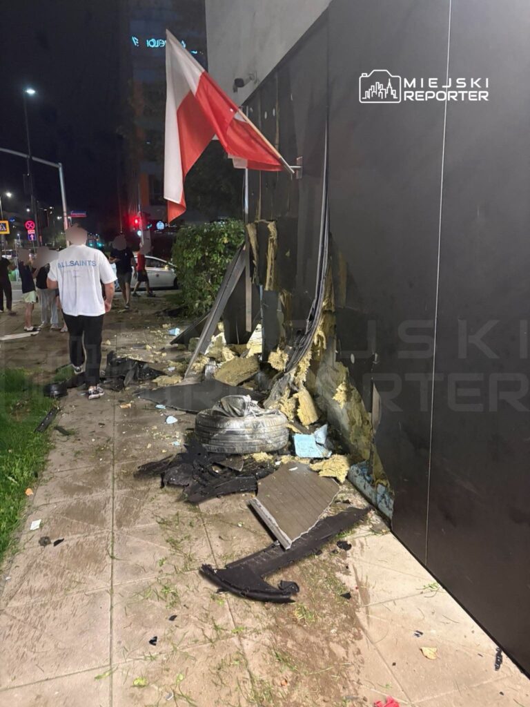 Grupa ludzi obserwuje zniszczoną elewację budynku, obok powiewa flaga Polski, a na ziemi leżą fragmenty materiałów budowlanych.
