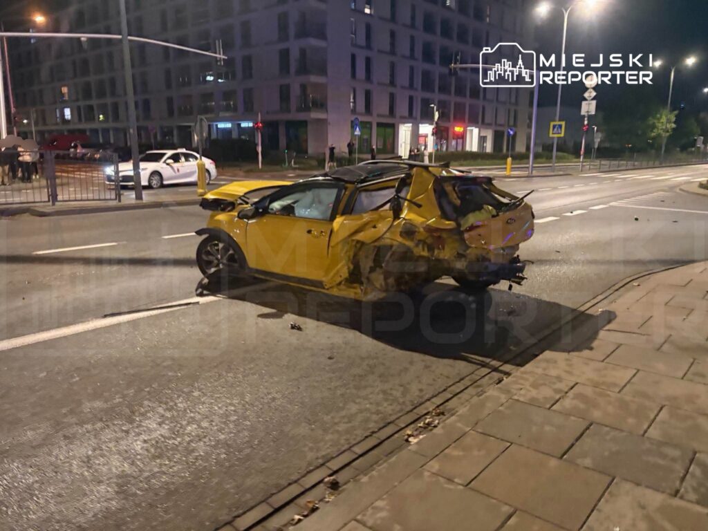 Zniszczony żółty samochód leży na jezdni po wypadku, otoczony przez inne pojazdy i przechodniów w nocy.