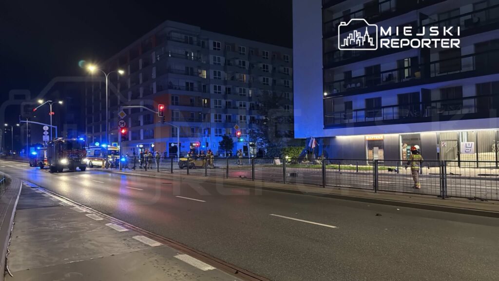 Na ulicy w nocy, strażacy i karetki interweniują przy nowoczesnym budynku mieszkalnym. W tle widać sygnalizację świetlną.