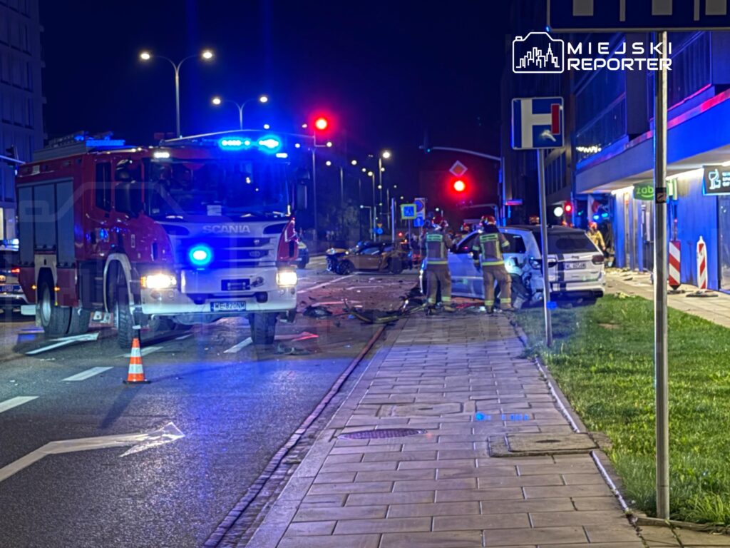 Czerwony wóz strażacki z włączonymi sygnałami świetlnymi stoi na drodze, a strażacy zabezpieczają miejsce wypadku.