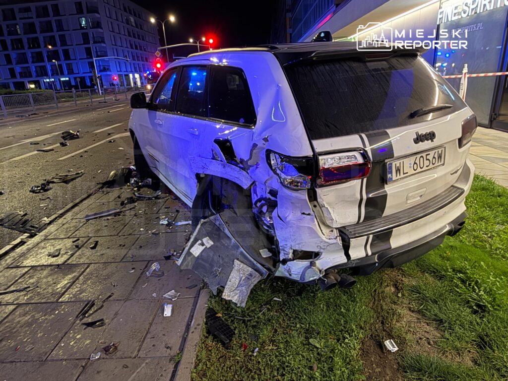 Uszkodzony biały SUV z widocznymi śladami wgniecenia stoi na poboczu, a wokół leżą fragmenty pojazdów.