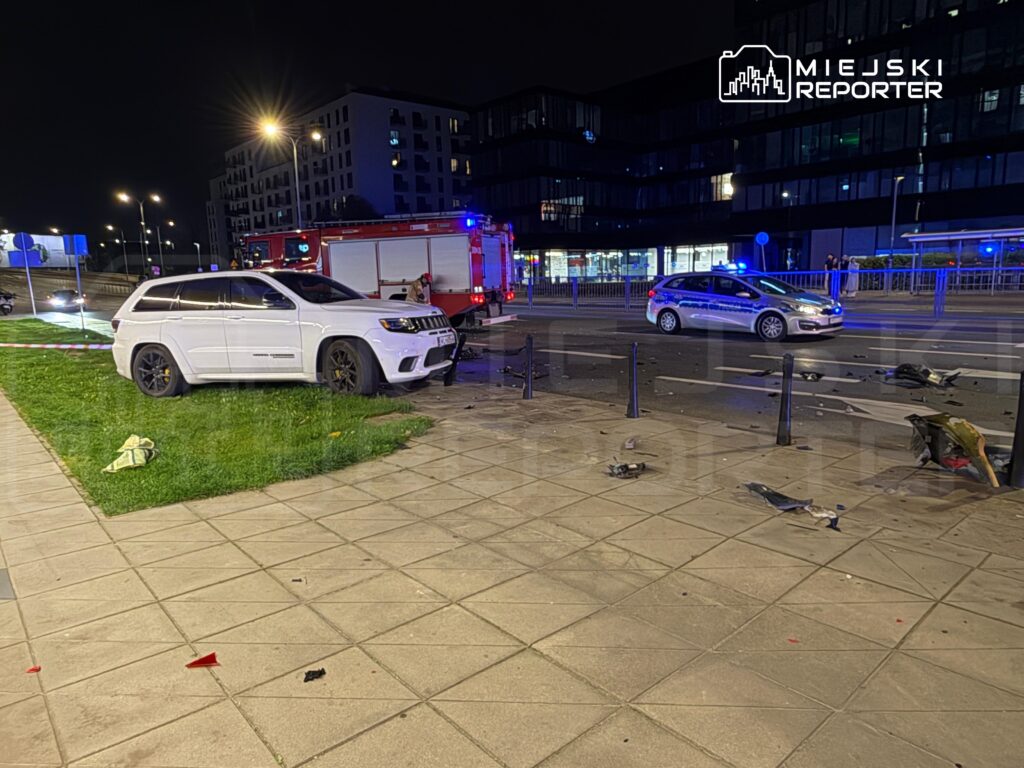 Biały SUV stoi na trawie po kolizji, obok widać uszkodzone elementy pojazdów oraz radiowóz z włączonymi sygnałami.