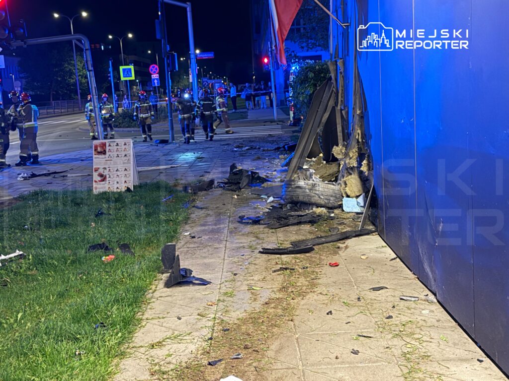 Grupa strażaków w mundurach na miejscu wypadku, w tle uszkodzona elewacja budynku i porozrzucane fragmenty.