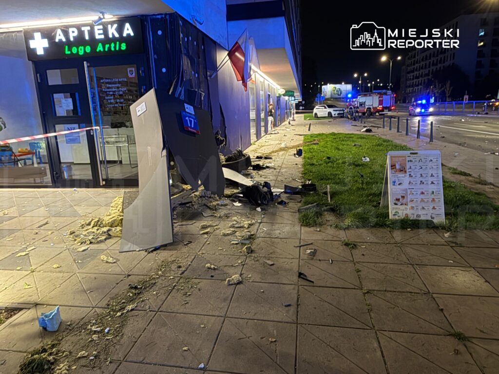 Zniszczona elewacja apteki po wypadku, z rozrzuconymi odłamkami i porozrzucanymi przedmiotami na chodniku. W tle widoczne pojazdy służb ratunkowych.