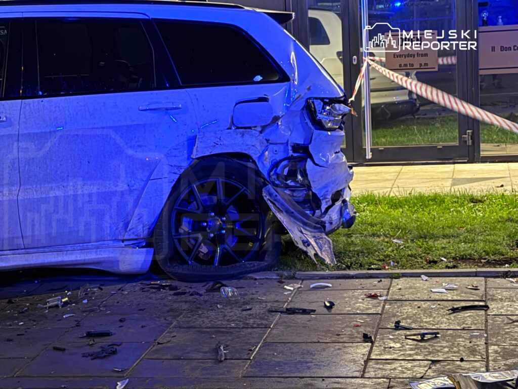 Uszkodzony biały SUV z wgniecioną tylną częścią stoi na chodniku, obok leżą fragmenty szkła i innych odpadków.