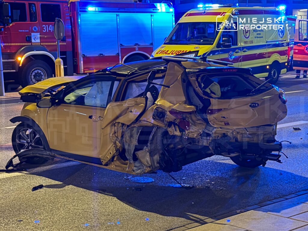 Zniszczony żółty samochód osobowy leży na drodze, obok niego stoją pojazdy służb ratunkowych z włączonymi sygnałami.