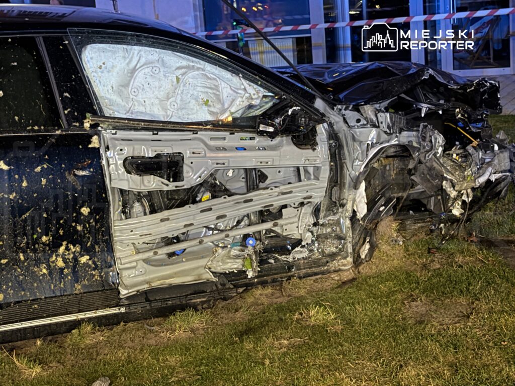 Zniszczony samochód leży na trawie, z uszkodzoną boczną częścią i otwartymi drzwiami.