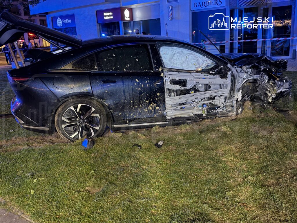 Zniszczony samochód osobowy leży na trawie obok budynku, z wygiętą karoserią i otwartymi drzwiami.