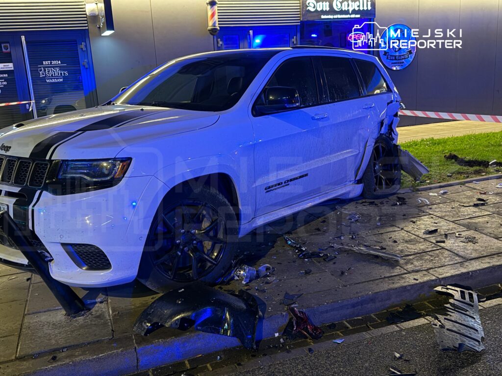 Biały SUV z uszkodzeniami tylnym zderzaku stoi na chodniku obok baru, wokół rozrzucone fragmenty karoserii.