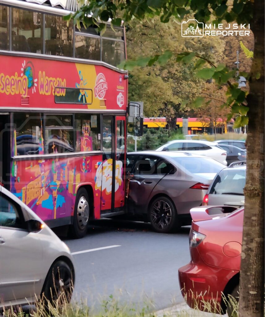 Czerwony autobus turystyczny z grafiką zderza się z szarym samochodem osobowym na ulicy.