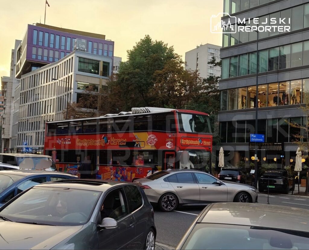Czerwony autobus turystyczny zatrzymany na ulicy wśród zaparkowanych samochodów, w tle nowoczesne budynki.