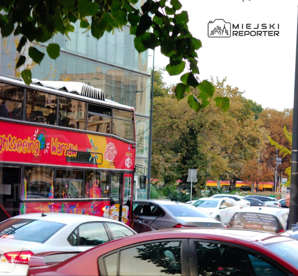 Czerwony autobus turystyczny z napisem "Sightseeing Warsaw" zatrzymany w ruchu ulicznym w mieście, otoczony samochodami.